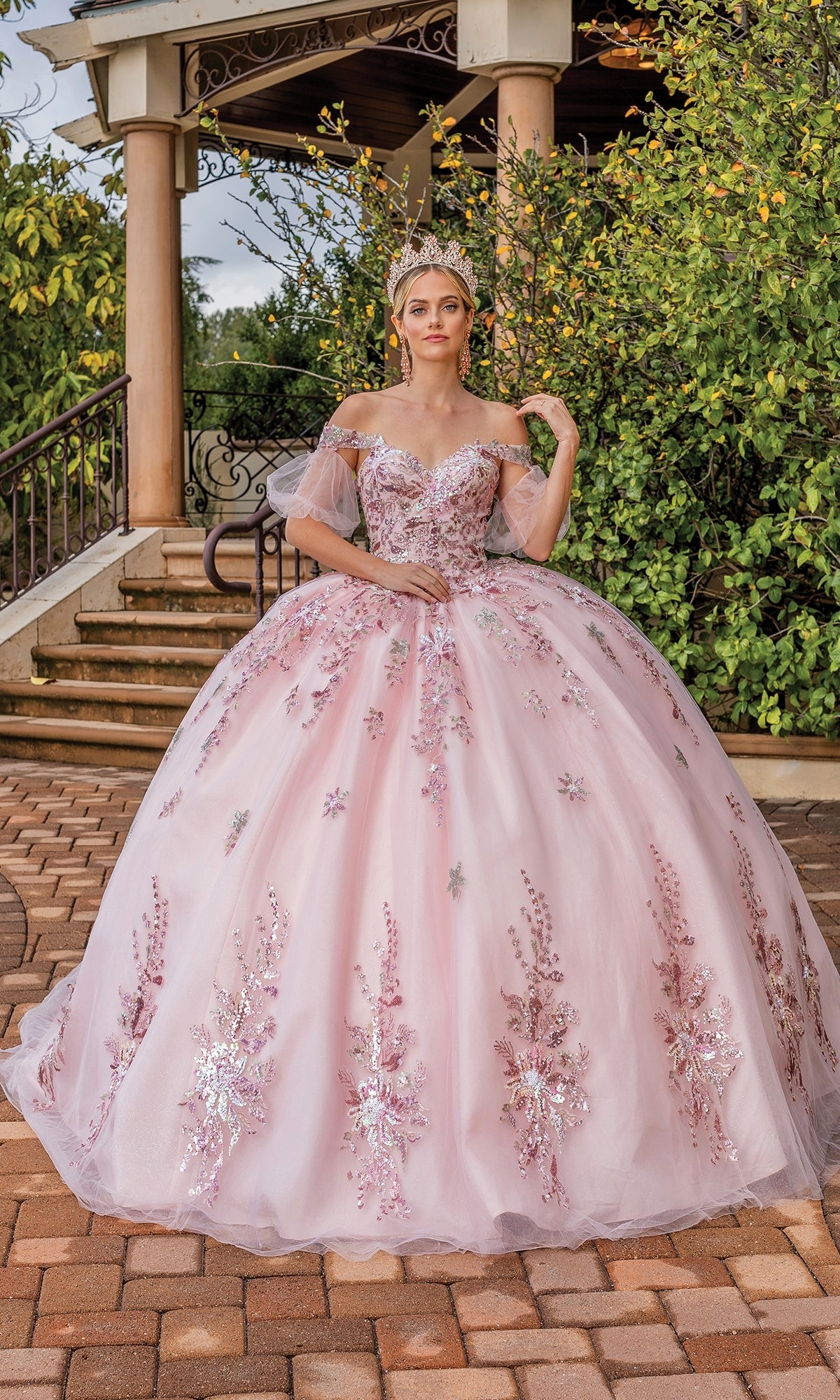 A woman stands outdoors by a staircase and greenery in the Quinceanera Dress 1821 By Dancing Queen, a blush pink ball gown with a sweetheart neckline, off-the-shoulder floral embellishments, and a tiara for an enchanting look.